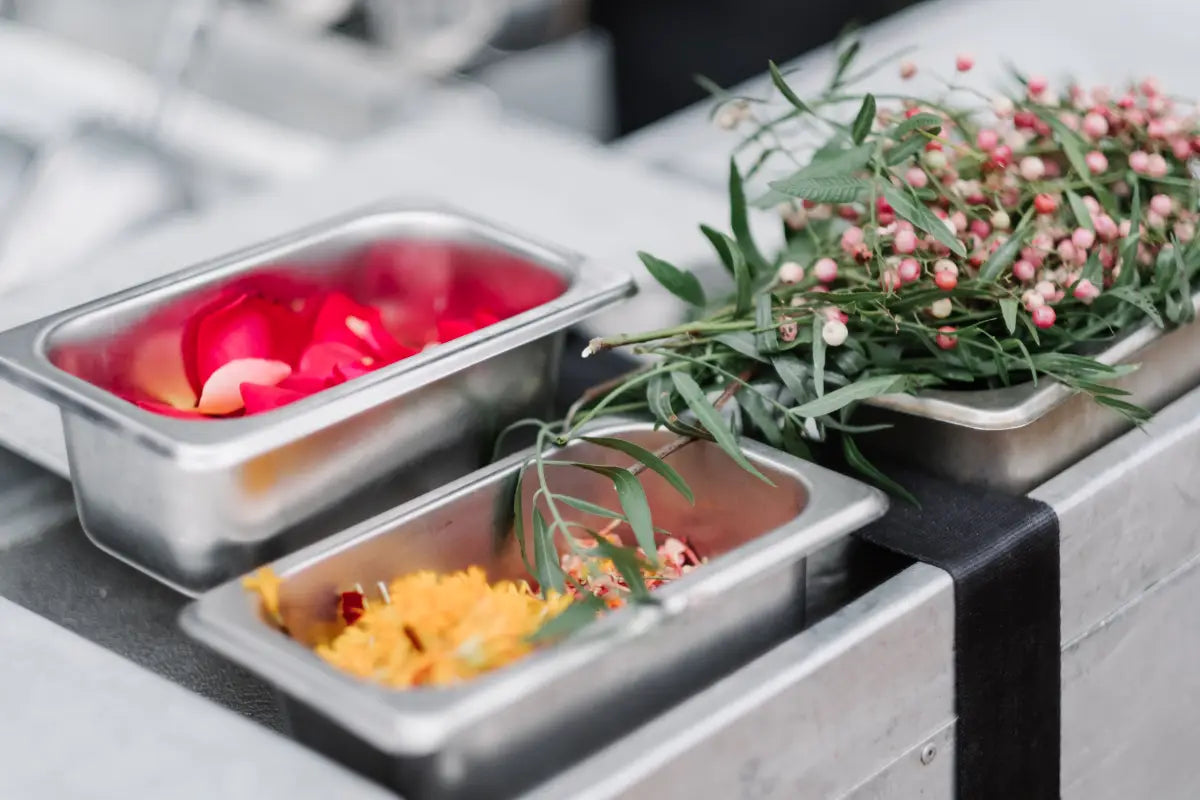 Fresh pink peppercorn branches and edible flower petals displayed in metal containers, ready for use as sustainable cocktail garnishes