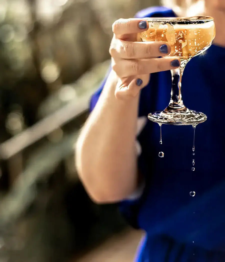 A clear, elegant champagne coupe glass filled with a golden-hued beverage and ice cubes.