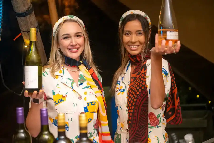 A clear glass bottle of rosé wine with a white label and gold foil cap, held up by one woman.