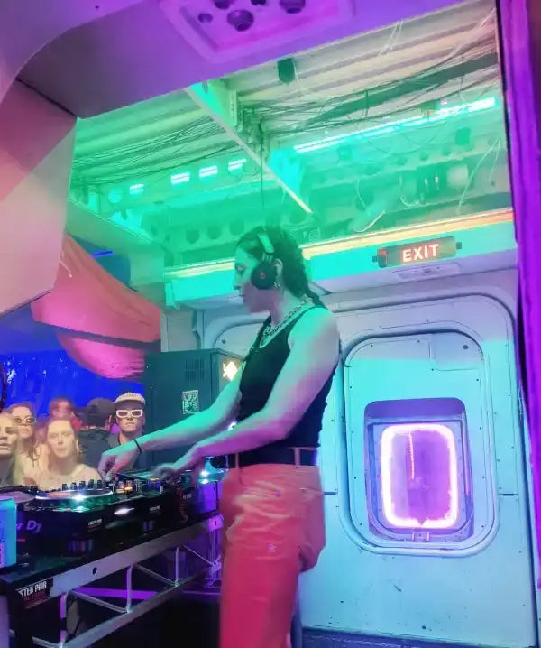 A dj in a black tank top and orange pants stands behind a professional audio mixing console, illuminated by vibrant purple and green stage lights.