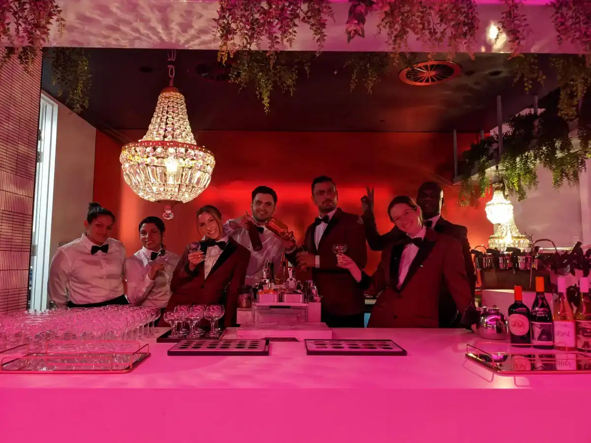 A group of bartenders in matching maroon tuxedos stand behind a glowing pink bar counter, holding glasses and bottles under the light of an ornate crystal chandelier.