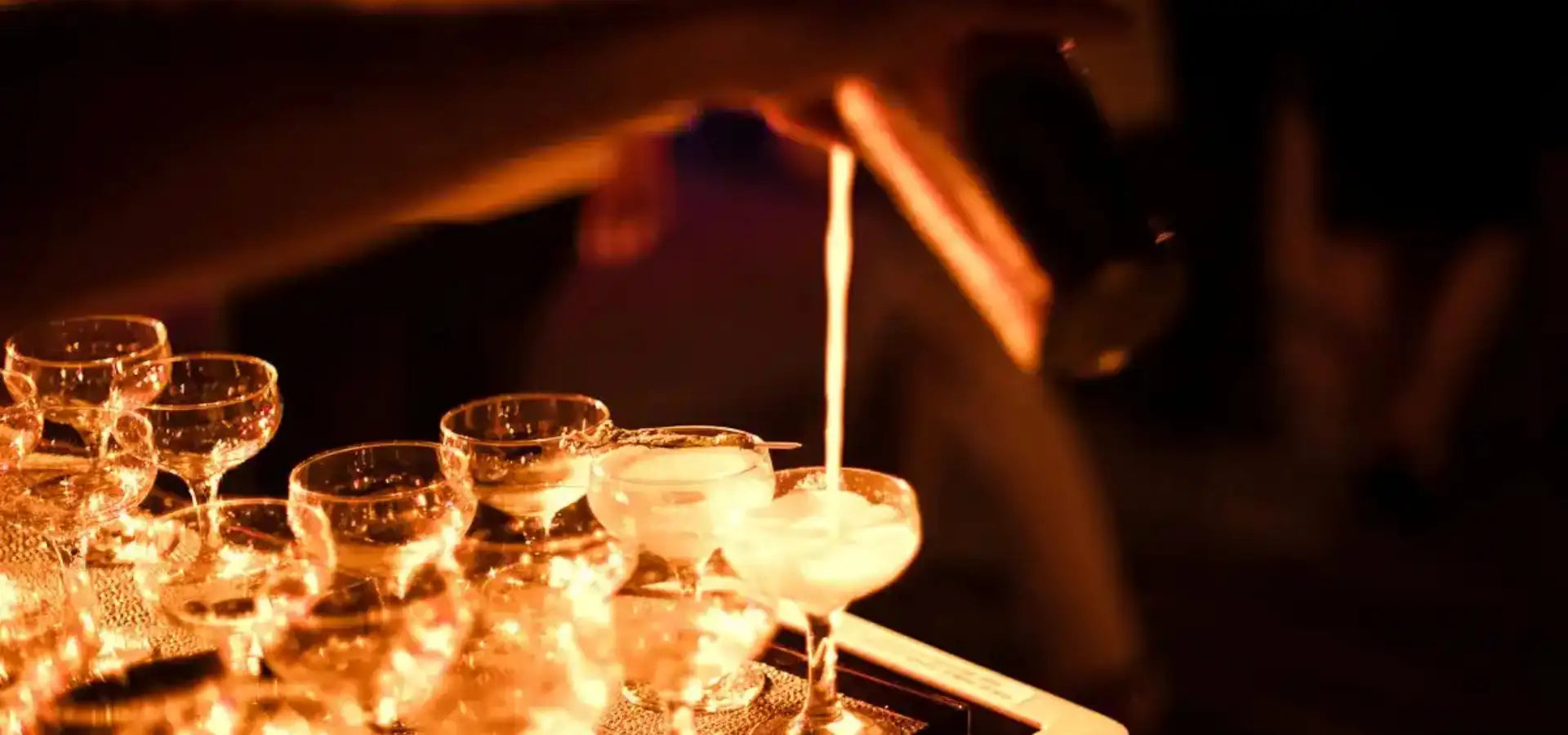 A hand pours a creamy white liquid into a clear stemmed cocktail glass, illuminating the scene with warm golden light.