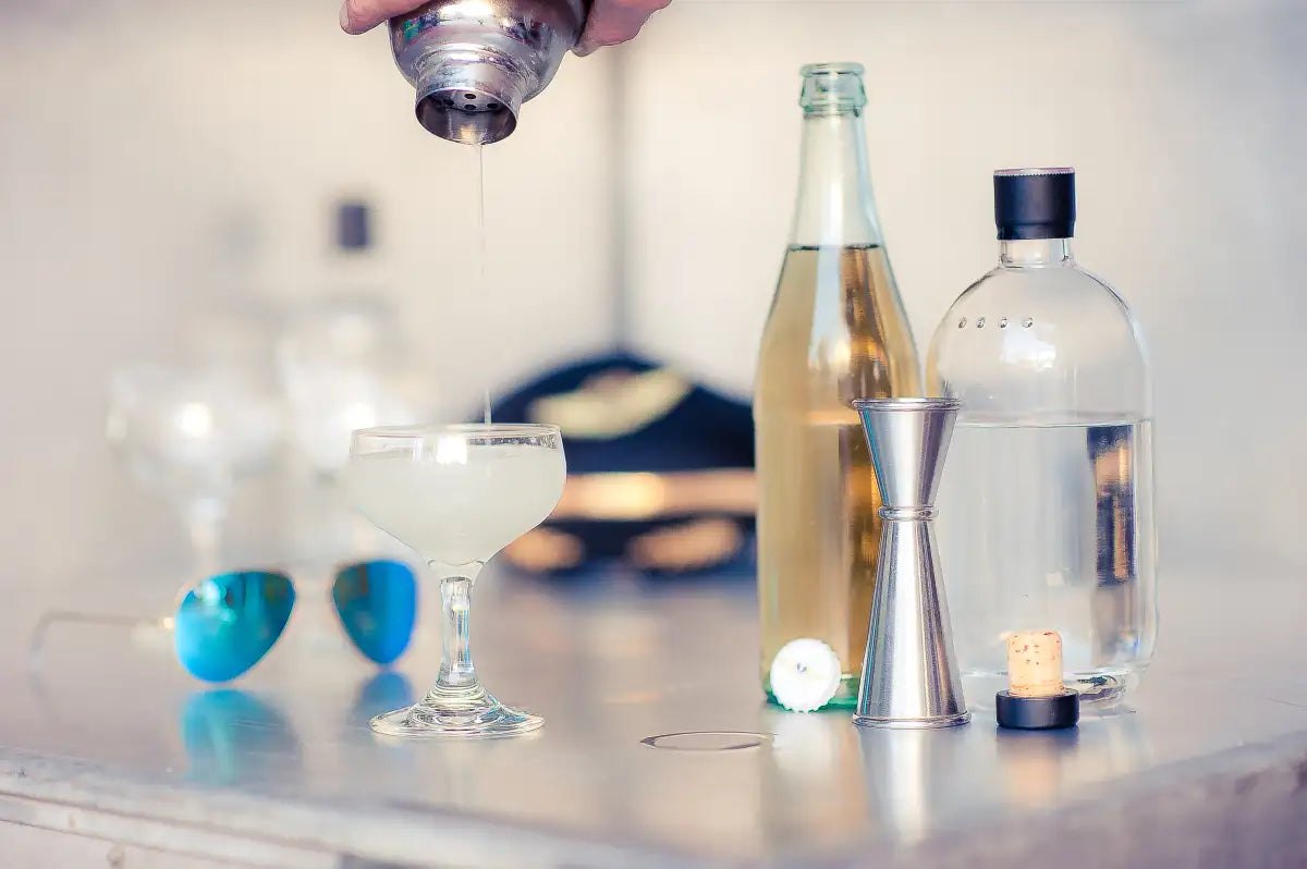 Cocktail being poured from a shaker into a coupe glass with aviation sunglasses, pilot cap, and bar tools in the background – Trolley’d cocktail class experience