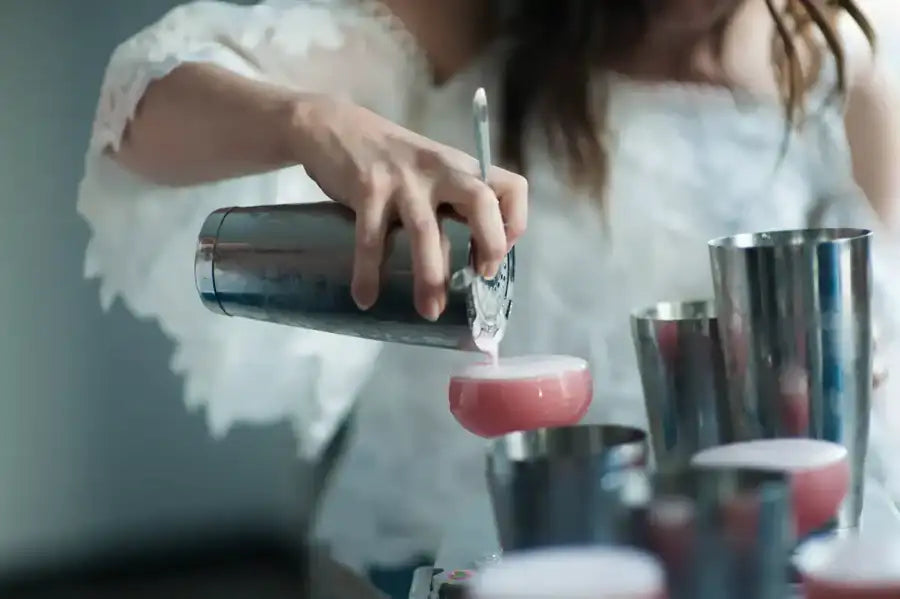 A sleek, stainless steel cocktail shaker being used to pour a pink drink into a coupe glass.