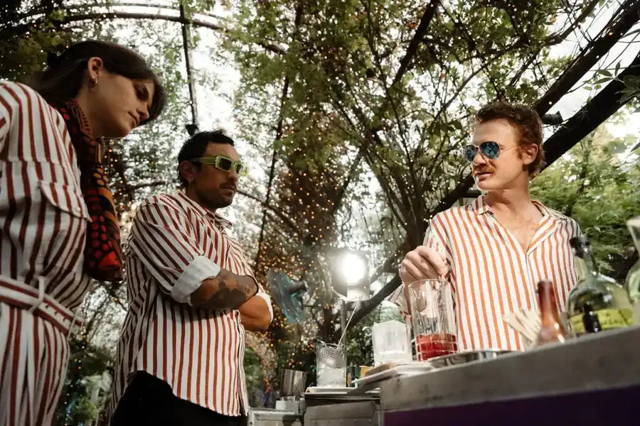 Three men in matching red and white striped shirts stand at an outdoor bar, one wearing green sunglasses while another holds a cocktail shaker.