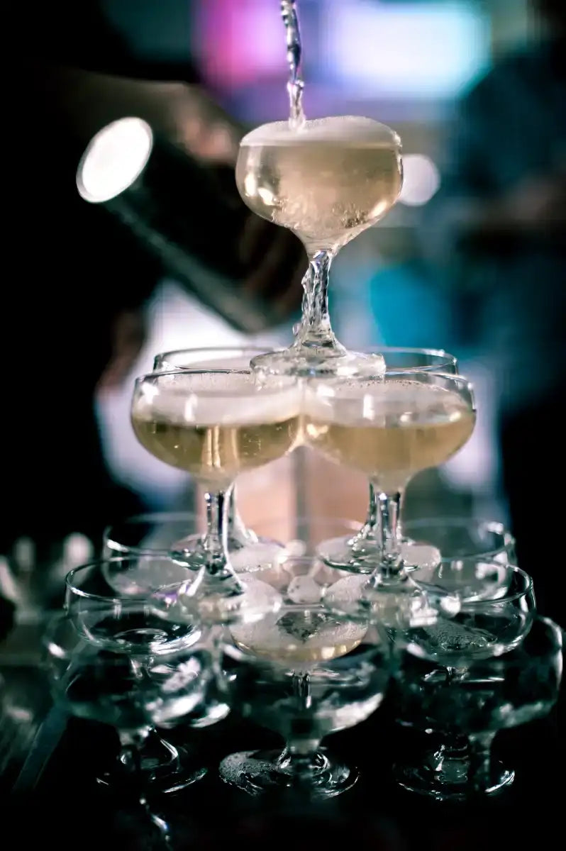 A tiered champagne pyramid with clear glass coupe glasses filled with golden bubbly liquid, topped by a pouring stream of sparkling wine.