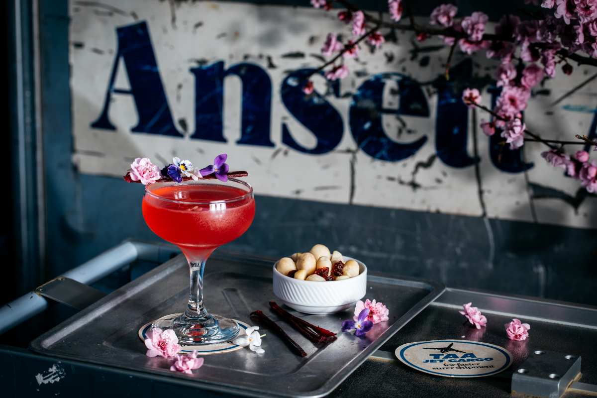 Vibrant pink cocktail garnished with edible flowers beside Ansett vintage airline signage, styled with cherry blossoms – Trolley’d signature drink