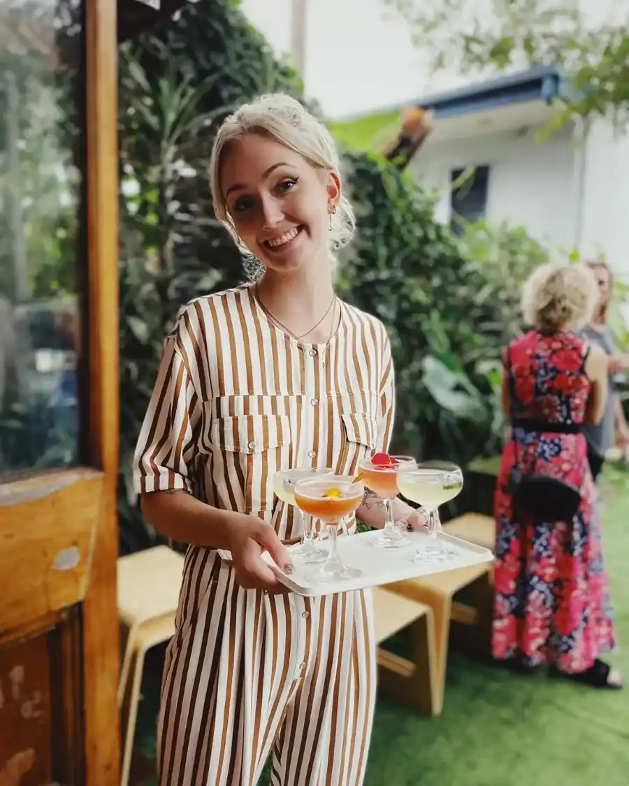 A woman in a beige and brown striped jumpsuit holds a white rectangular tray with three cocktail glasses.