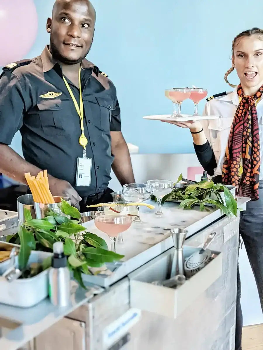 A woman in a white blouse and patterned scarf holds a silver tray with two pink cocktails.