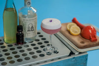 Pink Magma cocktail in a coupette glass with a frothy top and a violet garnish, surrounded by ingredients including lemon myrtle syrup, North Coast Bar Series rum, and Illawarra plum tincture.