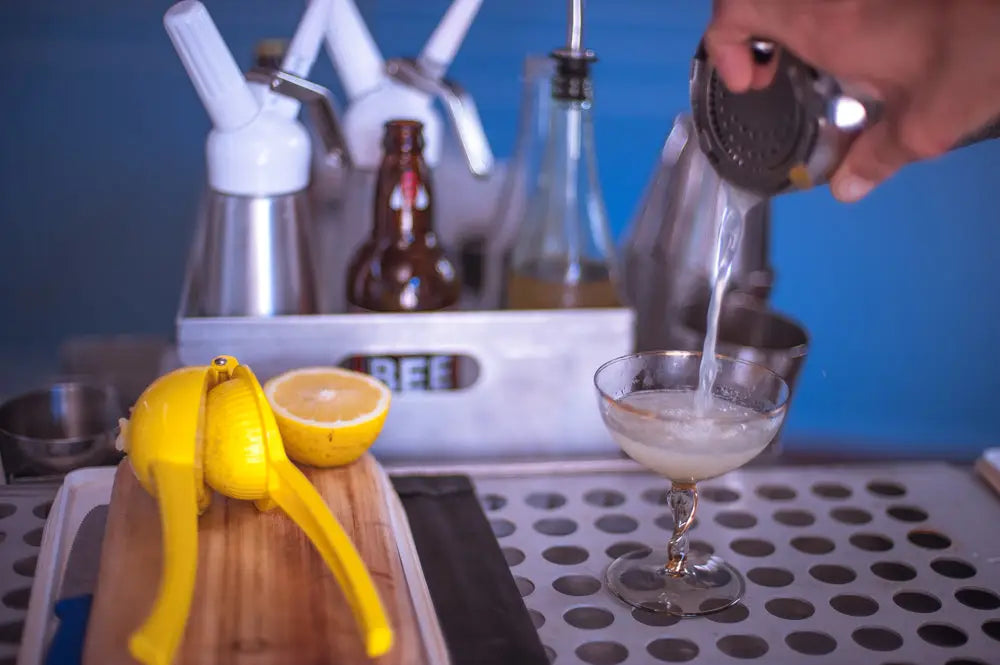 Bartender pouring infused tequila and lemon mix into a coupe glass for the Laughing Margarita, with a fresh lemon and bar tools in the background.