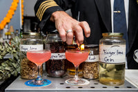 A Trolley'd bartender in a pilot uniform prepares vibrant pink cocktails using finger lime and pepper berry botanicals, with jars of ingredients labeled 'Finger Lime,' 'Pepper Berry,' and 'Finger Lime Gin' in the background