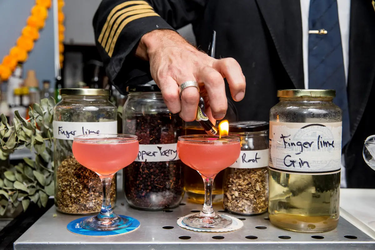 A Trolley'd bartender in a pilot uniform prepares vibrant pink cocktails using finger lime and pepper berry botanicals, with jars of ingredients labeled 'Finger Lime,' 'Pepper Berry,' and 'Finger Lime Gin' in the background