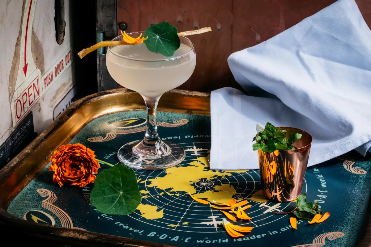 A refreshing River Mint, Mintbush & Blue Gum Daiquiri served in a coupe glass, garnished with dehydrated lemon and nasturtium leaf, on an aviation-themed tray with marigold petals.
