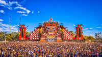 The iconic Defqon 2018 main stage featuring a striking design, vibrant lighting, and a massive crowd of festival-goers under a bright blue sky.