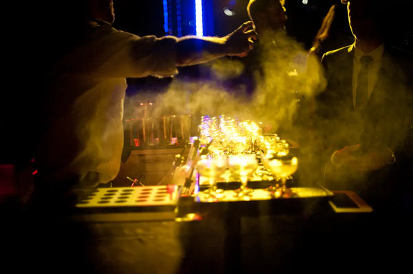 Atmospheric view of a bartender preparing cocktails amidst a cloud of mist at the Australian Bartender Awards 2023, with warm lighting illuminating the scene.