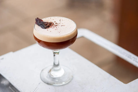 Close-up of a creamy Espresso Martini enhanced with Trolley'd Roast Wattle, Cacao & Honey Syrup, garnished with a delicate piece of dried fruit and a dusting of cacao, presented on an airline trolley side tray..