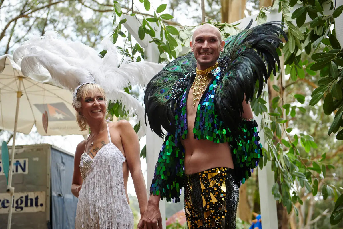 JP and PG, dressed in stunning feathered costumes, smile joyfully during their wedding ceremony at Wild Ducks in the Southern Highlands. The lush greenery and unique attire highlight the vibrant and personalized nature of their special day.