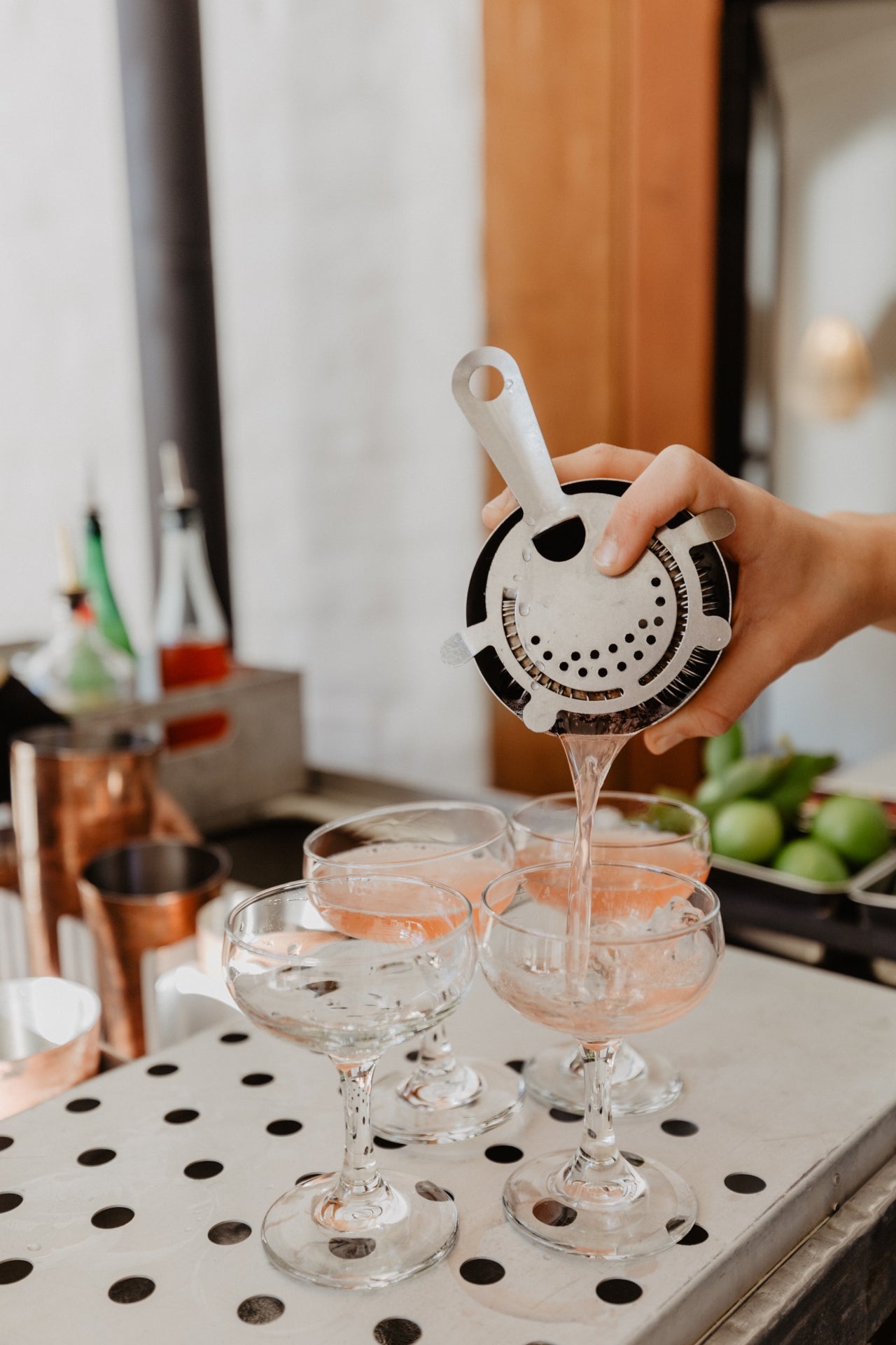 Bartender straining a cocktail into coupe glasses using a Hawthorn strainer during a hosted virtual cocktail class.