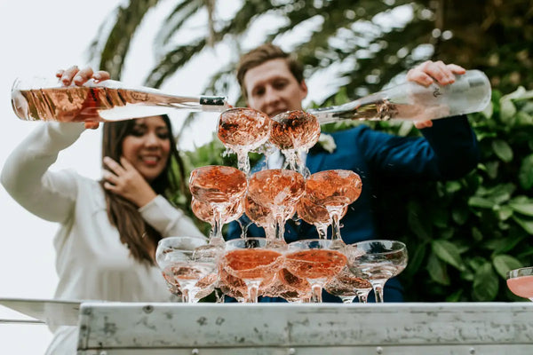 Joyous moment captured at an outdoor event where a man and woman pour rose champagne into a sparkling tower of glasses, arranged on a Trolley’d mobile bar. The scene is set against a lush green backdrop, highlighting the elegance and festivity of the occasion.