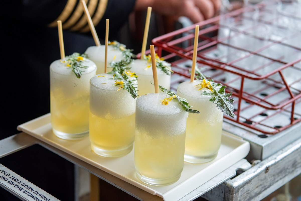 A tray of frothy, yellow dandelion cocktails garnished with green leaves and yellow petals, crafted by Trolley'd for a sustainable and eco-friendly drinking experience.