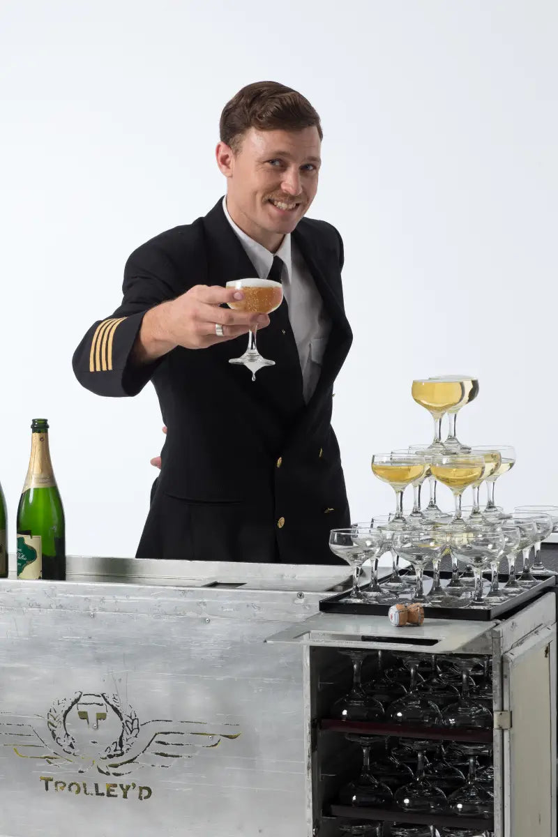 A Trolley'd bartender dressed in a pilot uniform serves champagne from a coupe glass at an elegant setup, featuring a champagne tower, showcasing Trolley'd's sophisticated glassware hire services.