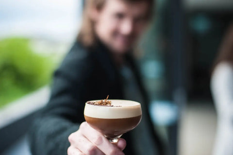Smiling person extending a hand to offer a Trolley'd Espresso Martini, topped with a sprinkle of cacao and garnished with a unique botanical, embodying the art of modern mixology