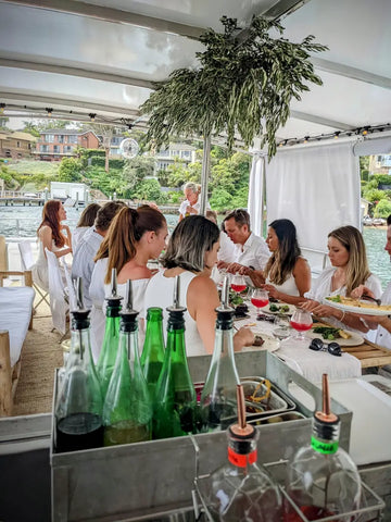 A group of friends in white outfits enjoying drinks and food at an elegant outdoor dining setup provided by Trolley’d.