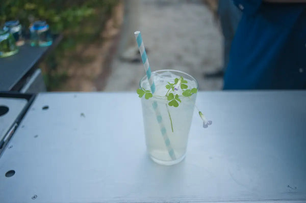 A glass of Lemon Myrtle Lemonade mocktail garnished with clover leaves and flowers, served with a blue and white striped straw on a bar counter at a Trolley'd mobile bar setup.