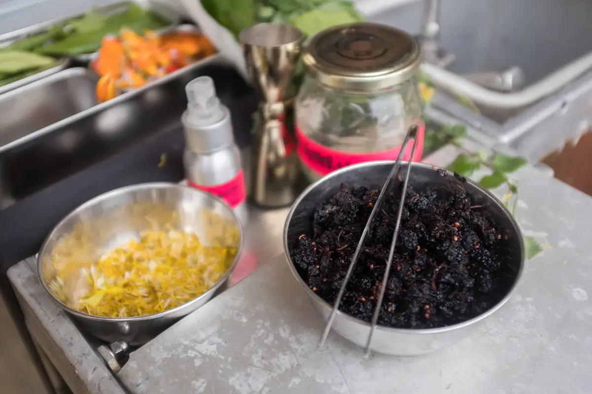 A close-up of foraged mulberries and yellow native blossoms in metal bowls on the Trolley’d bar, surrounded by fresh herbs and house infusions — showcasing the craft of sustainable cocktail preparation.