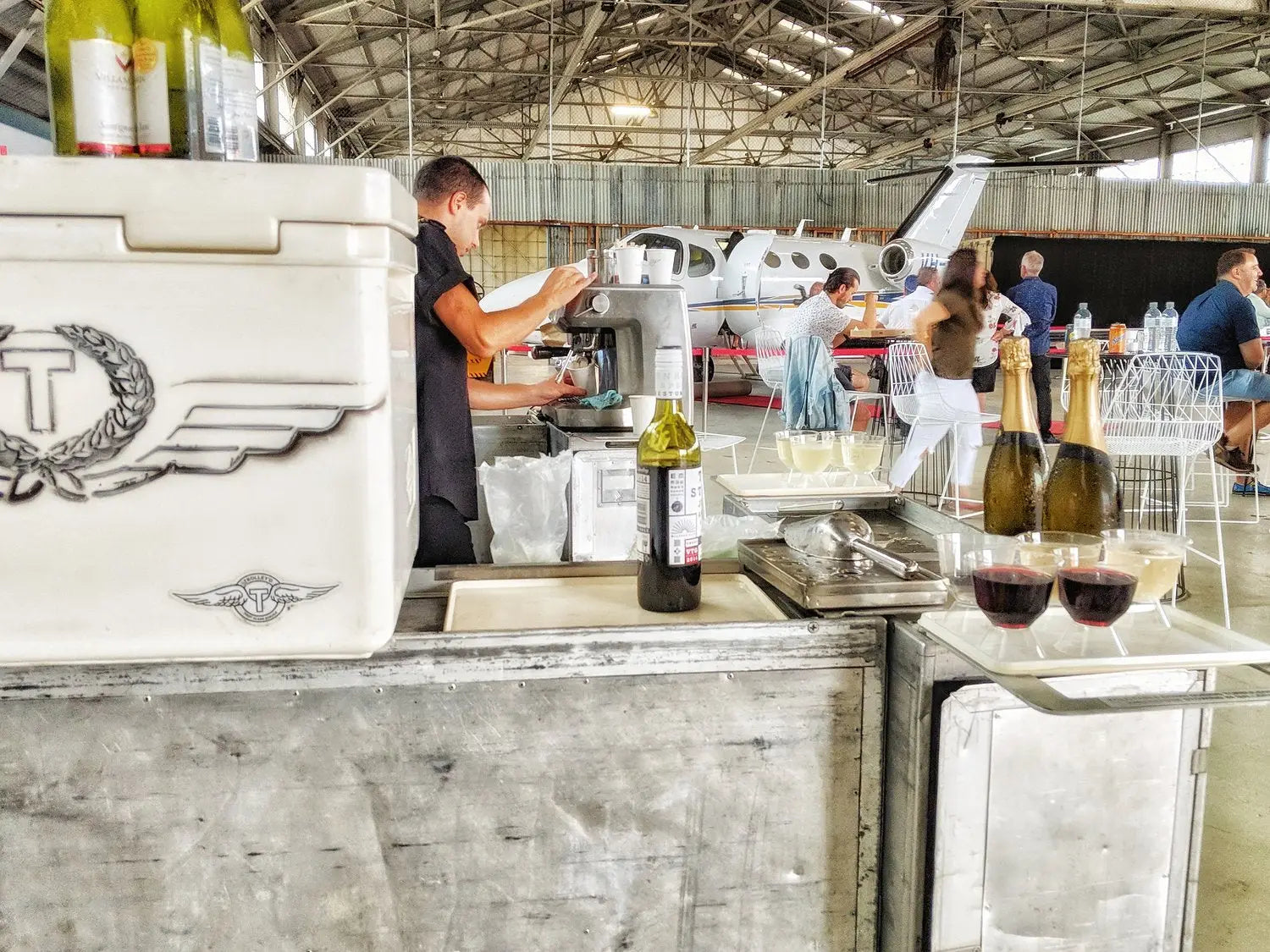 Busy event scene featuring Trolley'd mobile bar and coffee van setup in an airplane hangar, with a bartender preparing drinks. The bar displays an assortment of beverages including wine, champagne, and crafted cocktails, set against the backdrop of a small private jet and a gathering of guests, creating a dynamic and upscale event atmosphere.