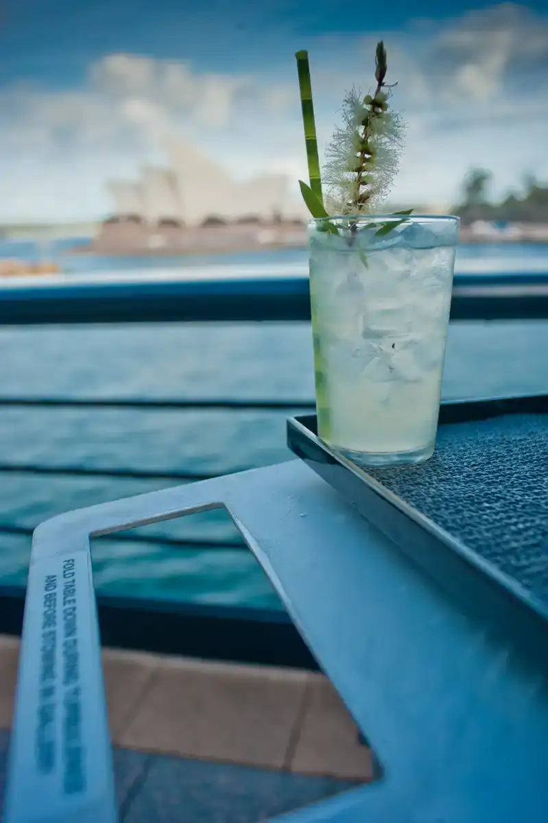 A clear glass tumbler filled with a light green cocktail and ice, garnished with a bamboo straw and a delicate white flowering plant.