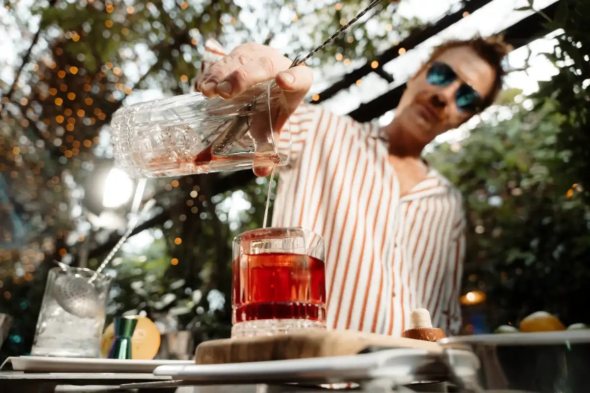 A bartender in sunglasses and a striped shirt pours a deep red Negroni from a crystal mixing glass into a tumbler with a large ice cube. The setting is an outdoor bar with warm string lights and lush greenery, creating an elegant and immersive cocktail experience.
