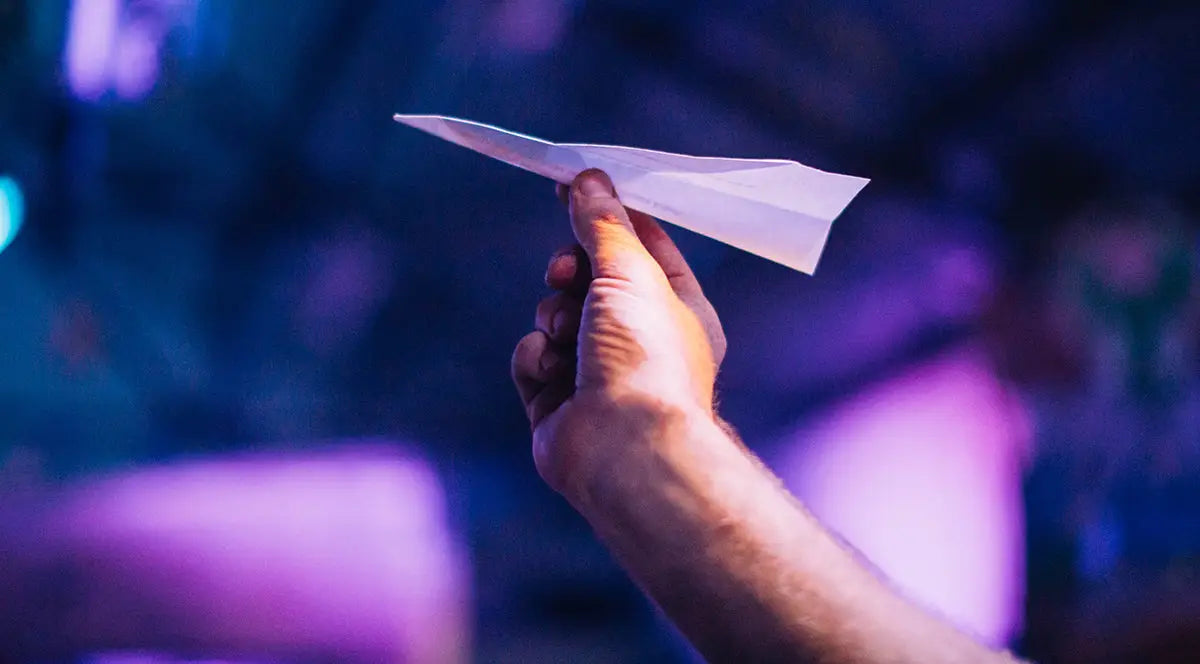 A close-up shot of a hand confidently launching a paper airplane, set against a blurred background illuminated with purple and blue lighting, symbolizing the playful and imaginative spirit of events catered by Trolley'd Mobile Bar.