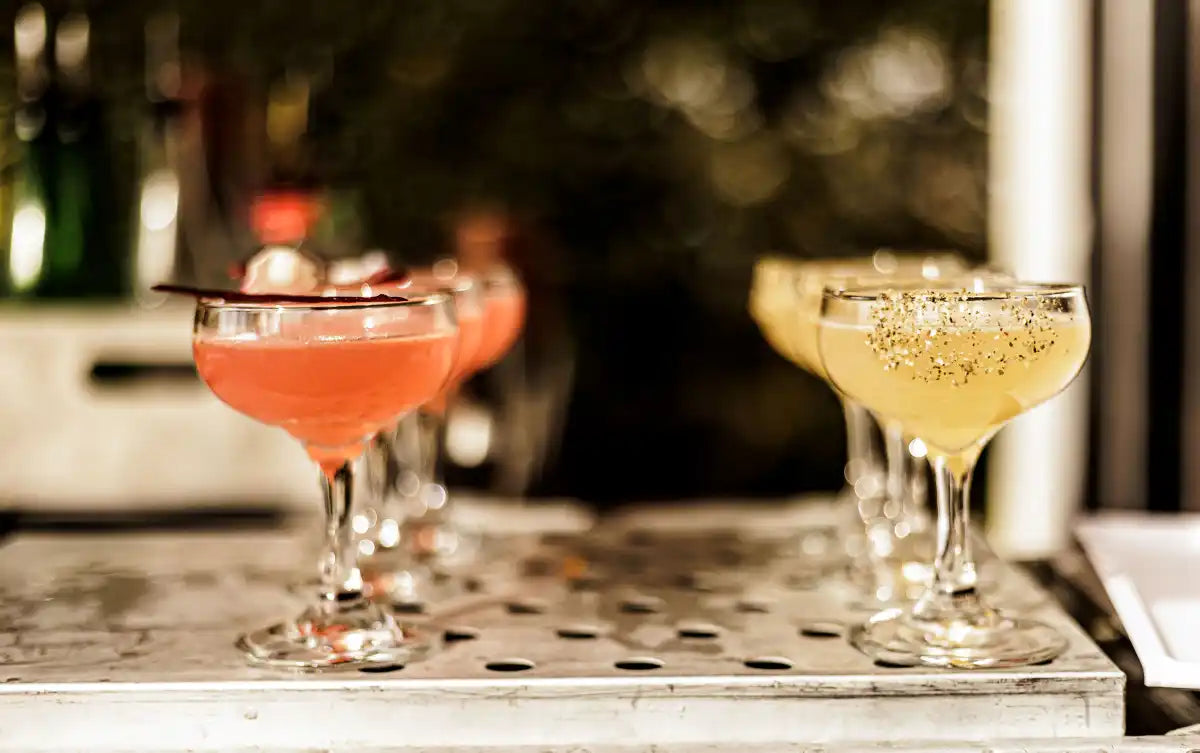 Elegant coupe glasses filled with vibrant cocktails, one pink with a garnish and the other a golden drink with a decorative rim, prepared by Trolley'd mobile bar, placed on a metallic surface with a blurred background of a festive outdoor setting.