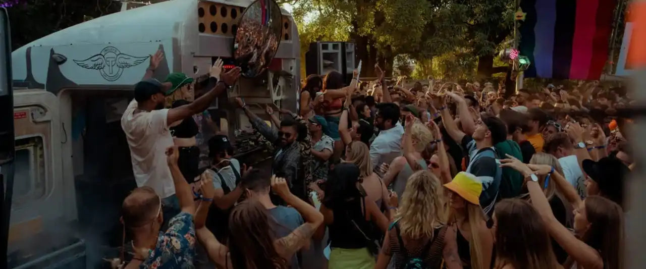 A lively festival crowd enjoying the music and atmosphere in front of the Trolley'd mobile bar, with people dancing and raising their hands. The Trolley'd logo is visible on the bar, surrounded by trees and colorful festival decorations.