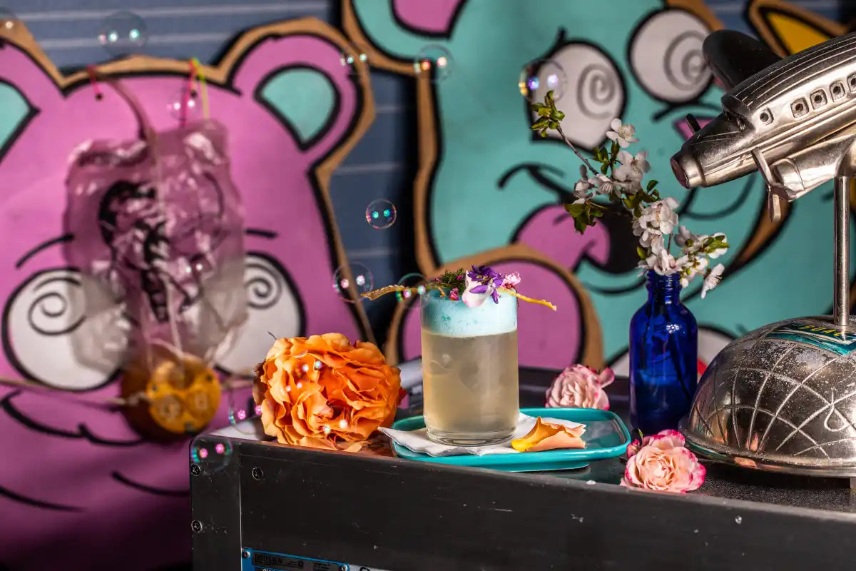 A handcrafted cocktail from Trolley'd featuring a frothy top, garnished with vibrant flowers and herbs, set against a whimsical background with colorful bear illustrations and aviation-themed decor, including a vintage airplane model and bubble effects.