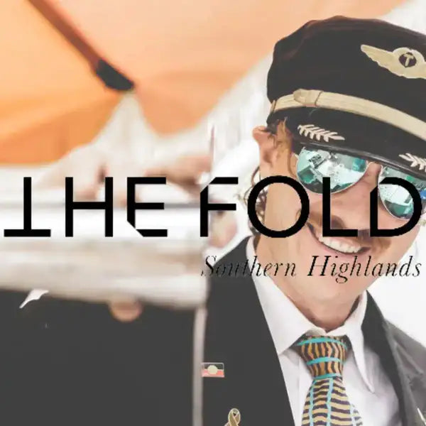 A smiling man wearing a black pilot cap with gold wing and laurel emblem, reflective blue-tinted sunglasses, and a patterned tie.