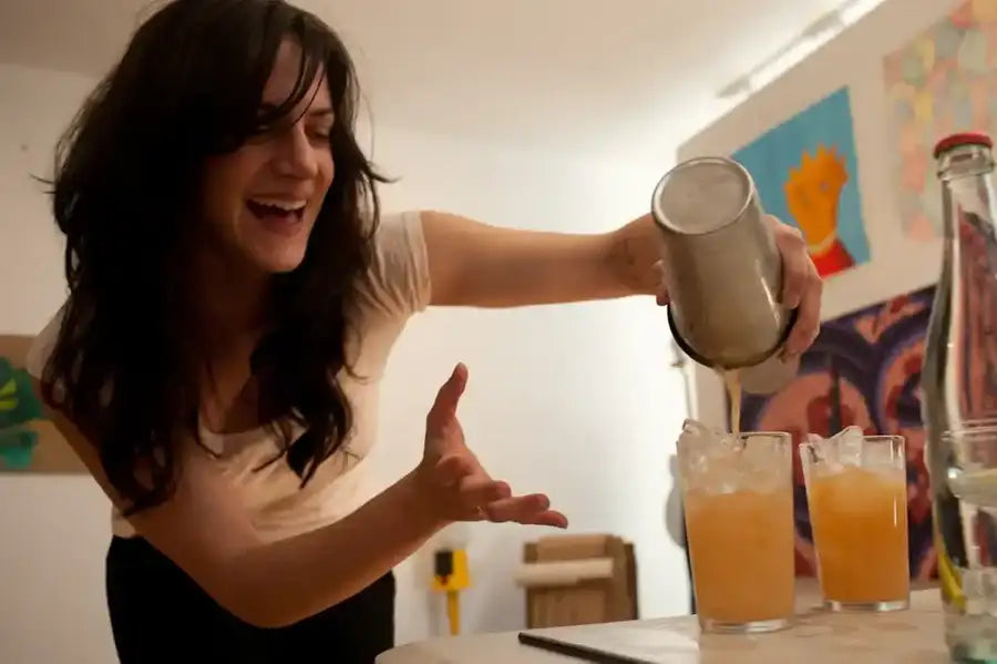 A metallic cocktail shaker being used to pour a golden-orange liquid into glasses.