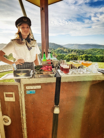 A woman dressed in a vintage flight attendant uniform stands behind a mobile bar adorned with an array of colorful cocktails and fresh garnishes. The bar has a rustic charm, featuring a copper top and a wooden structure with visible rivets, reminiscent of an old-fashioned steamer trunk. The setting is outdoors with a beautiful, lush green mountain landscape in the background under a clear blue sky, suggesting an upscale event in a picturesque location. The mobile bar carries a tag labeled "ANSETT AUSTRALIA ANI 928," adding to the overall vintage aviation theme of the setup.