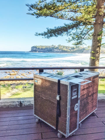 A rustic mobile bar with a copper countertop is situated on a wooden deck overlooking a scenic beachfront. The bar, resembling a vintage trunk on casters, has a copper sheet finish with metal reinforcements and rivets. The backdrop features a picturesque view of Palm Beach with waves gently crashing near the shore, and a lush pine tree framing the left side of the image, enhancing the natural beauty of the location. The setting is serene and ideal for a beachside event or gathering where a mobile bar can provide refreshment while guests take in the stunning coastal views.