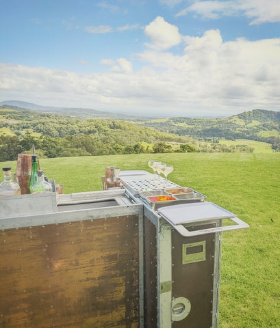 A mobile bar cart with a copper top is stationed outdoors on a hill, with a panoramic view of rolling green hills and a distant coastline under a partly cloudy sky. The bar cart is made of a weathered copper sheet with metal rivets and trim, giving it a vintage, industrial look. On top of the cart, there are several glass bottles of varying shapes, two elegant martini glasses with clear liquid, and a wooden holder containing garnishes, ready to serve. It presents a serene setting for an outdoor event where guests can enjoy crafted cocktails amidst the beauty of nature.