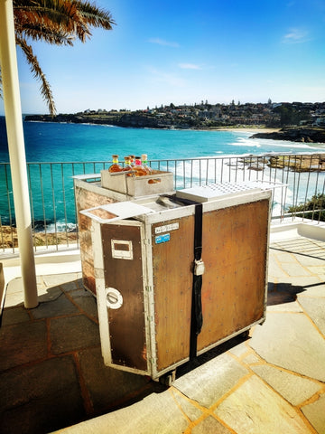 A copper-plated airline trolley-style mobile bar is positioned on a stone-tiled patio overlooking the stunning Tamarama Beach. The bar cart, featuring a copper sheet covered airline trolley with metallic corners and rivets, creates a vintage aesthetic. On top of the bar, there are several jars with colorful contents, ready for cocktail preparation. The location offers a breathtaking ocean view, with the coastline stretching into the distance, making it a perfect setting for an elegant outdoor event or a casual gathering by the sea.