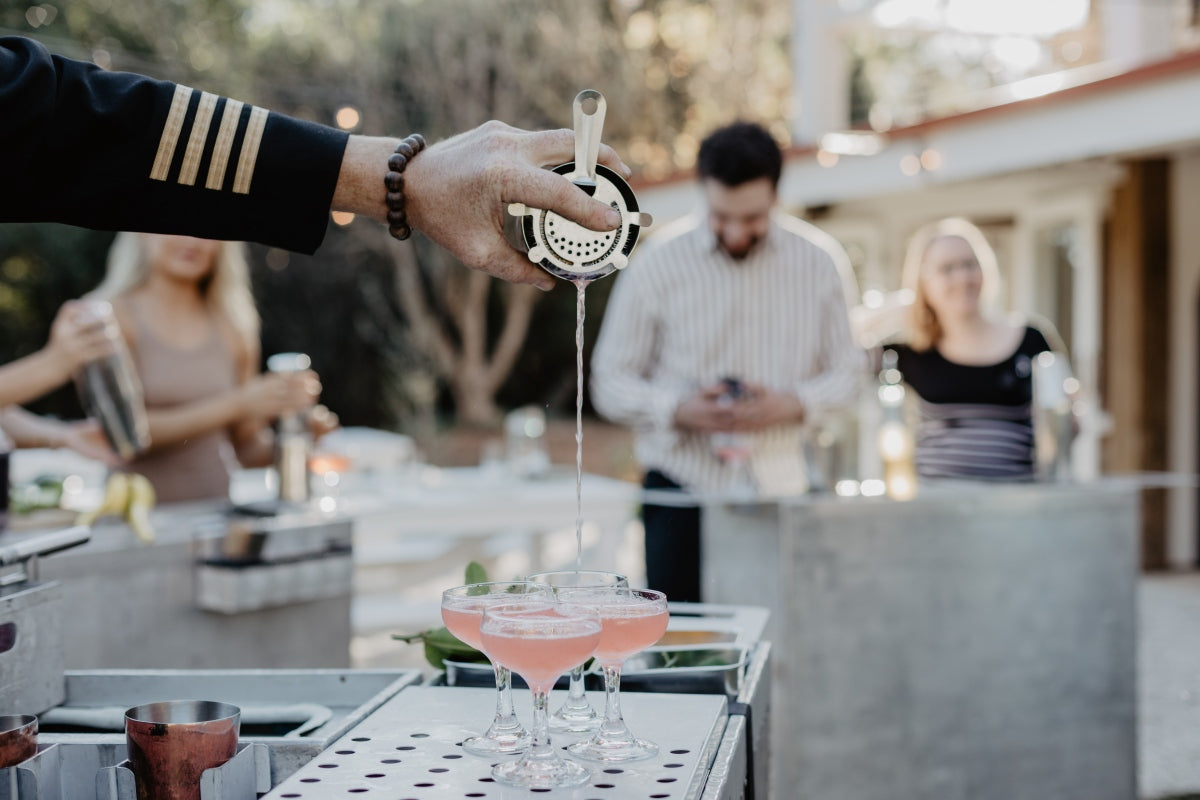 A Trolley’d captain pouring pink cocktails through a strainer, with class guests behind the airline trolley