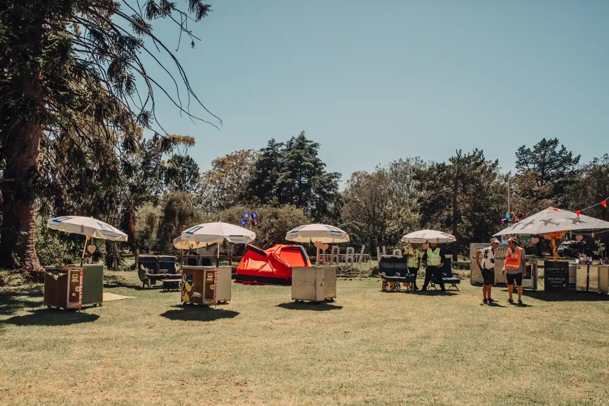 Aviation-themed outdoor event setup by Trolley'd featuring airline trolleys, umbrellas, and seating arrangements on a sunny day in a lush green park. Staff members in safety vests can be seen managing the setup.