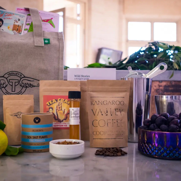 Image showcasing the contents of the Ultimate At-Home Cocktail Kit, including a Trolley'd hemp bag, Alto martini olives, Olsson’s sea salt flakes, Wild Stories foragers guide, Kangaroo Valley coffee, Tea Craft Egyptian iced tea, Urban Beehive honey, fresh lemons, and stainless steel cocktail tools.