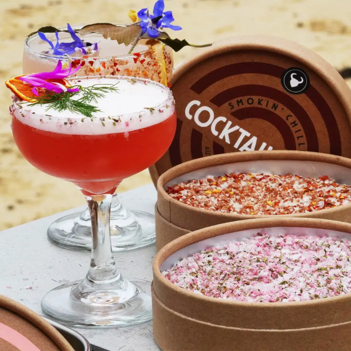 A selection of beautifully garnished cocktails featuring gourmet cocktail salts in various flavors. The cocktails are displayed on a beachside table, emphasizing the premium quality and visually appealing presentation of the drinks and salts.