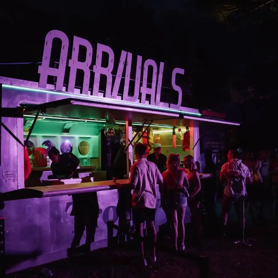 Purple neon-lit ’arrivals’ sign above a glowing green food truck.