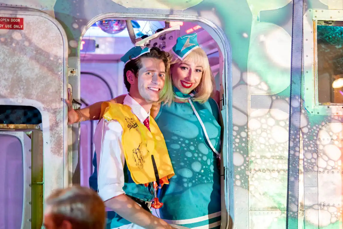 Trolley’d performers dressed as vintage flight attendants smiling at the doorway of a repurposed airplane installation at Neon Dreams, Vivid Sydney, lit with colourful bubble projections
