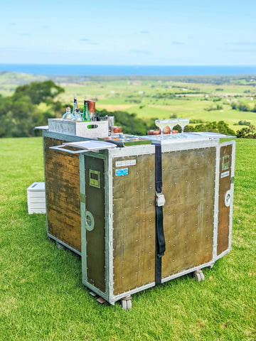 A mobile bar setup on a lush green hillside with a breathtaking view of the coastline in the distance. The bar, which resembles a vintage travel trunk on wheels, has a copper countertop and is constructed with a wooden frame accented with metal rivets. On top of the bar, there are several bottles of spirits and mixers, along with two copper mugs and a pair of cocktails ready to be enjoyed. The bar is tagged with "ANSETT AUSTRALIA ANI 928," giving it a unique, retro aesthetic. The scene suggests an elegant, outdoor event or gathering, offering guests not only refreshing beverages but also a stunning natural backdrop.